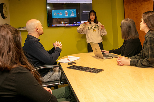 Avery stnading in front of a monitor delivering a presentation to Marketing Department staff members
