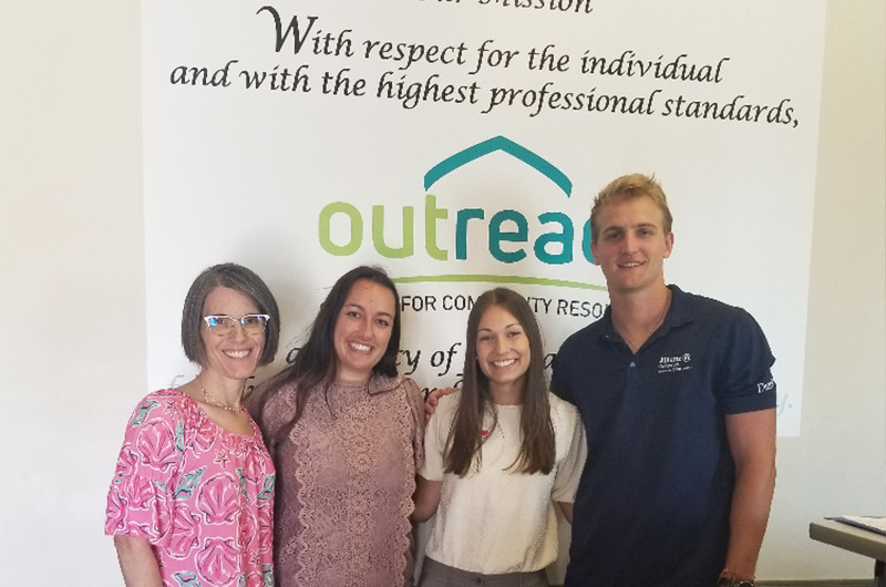 a woman professor standing next to three students, two women and one man, smiling in front of a backdrop with the outreach career fair logo for a posed photo