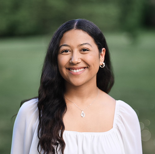 girl with long black hair in grass field smiling wearing a white shirt and silver necklace