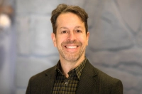 man smiling in a brown sport coat and shirt against a stone wall
