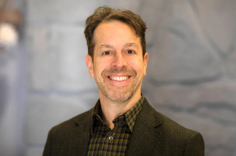 man smiling in a brown sport coat and shirt against a stone wall