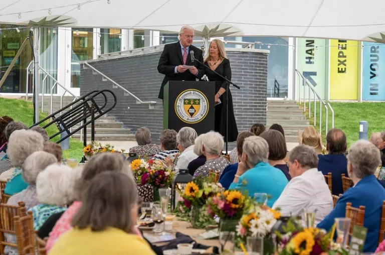 People at a podium in front of a crowd sitting at tables with flowers