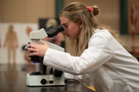 A female student in a white lab coat looks into a microscope.