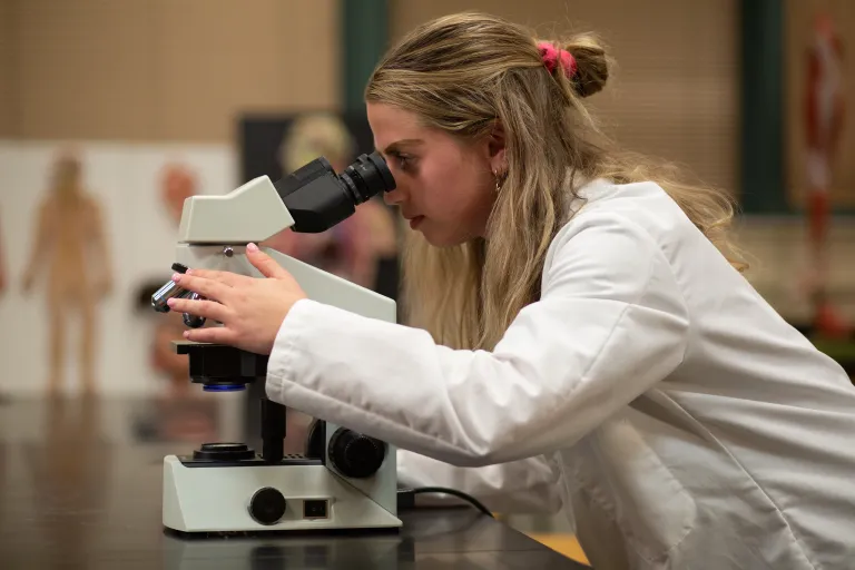 A female student in a white lab coat looks into a microscope.