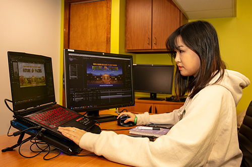 a side picture of Avery working at a desk with two computers in front of her