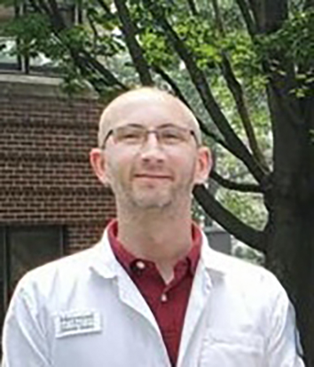 man posing in front of tree and brick buliding wearing white lab coat