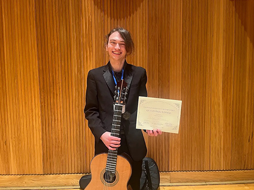 student standing in front of wooden wall with guitar and award in hands