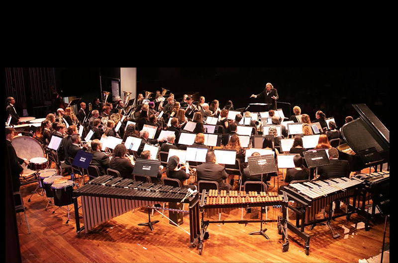 aerial picture from behind of the wind symphony performing and the conductor standing in front of them conducting
