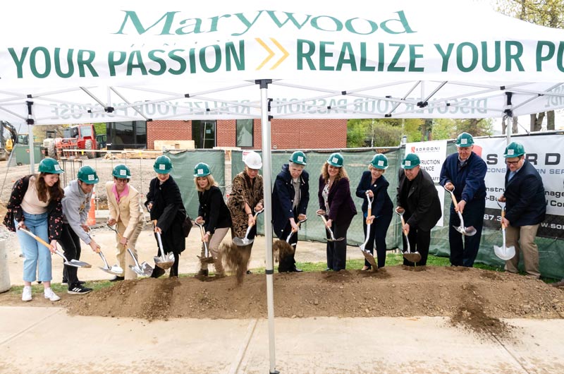 a group of people with shovels pretending to dig into the dirt for the groundbreaking of a new building wearing green hard hats with a white Marywood M on the front
